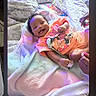 Gavianna'S is registered to the contest to win money with this photo: baby, bed, blanket, cartoon_clothing, child, closeup, family_photo, happy, home, indoor, infant, lying_down, messaging_app, orange_outfit, patterned_bedding, phone_screenshot, portrait, reaction_emojis, relaxed, smiling
