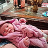 baby, infant, child, portrait, pink_blanket, bunny_hood, sleeping_baby, wide_eyes, indoors, living_room, coffee_table, baby_bottle, diaper, baby_wipes, fan, backpack, flowers, furniture, sitting, family_scene