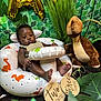 baby, infant, child, dinosaur, plush_toy, inflatable_chair, milestone_sign, greenery, leaves, plant, background, portrait, cute, indoor, sunglasses, decor, photography, smile, face, feet