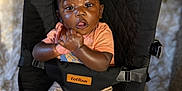 Asher is registered to the contest to win money with this photo: baby, child, infant, black_hair, orange_shirt, beige_shorts, brown_sandals, baby_seat, totrun, carpet, indoor, curious_expression, clasped_hands, sitting, cushion, footwear, person, portrait, young_child, comfortable