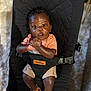 baby, child, infant, black_hair, orange_shirt, beige_shorts, brown_sandals, baby_seat, totrun, carpet, indoor, curious_expression, clasped_hands, sitting, cushion, footwear, person, portrait, young_child, comfortable