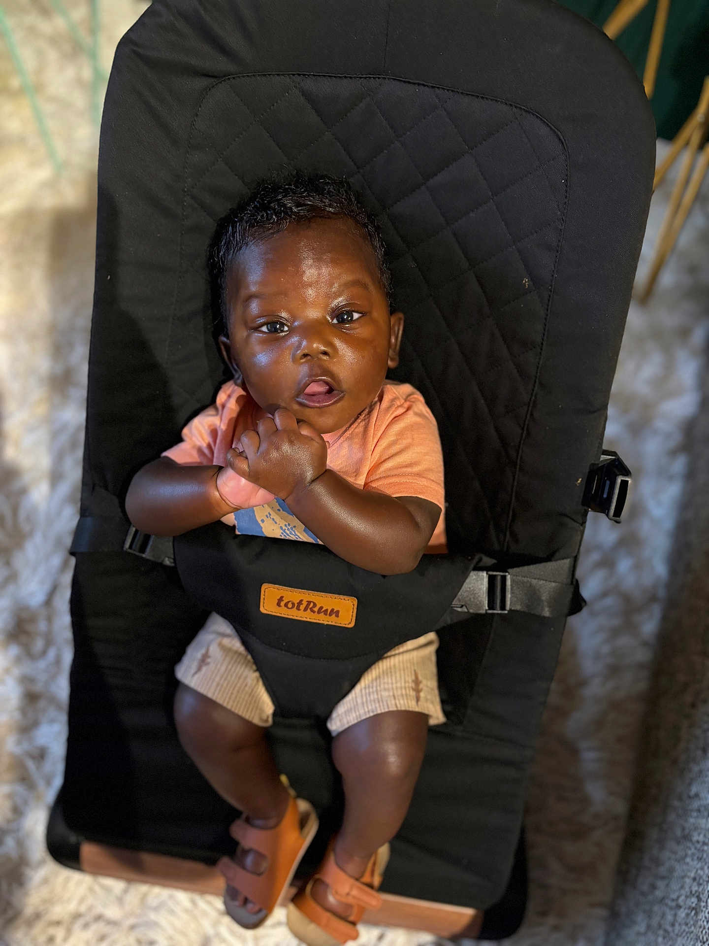 Asher is registered to the contest to win money with this photo: baby, child, infant, black_hair, orange_shirt, beige_shorts, brown_sandals, baby_seat, totrun, carpet, indoor, curious_expression, clasped_hands, sitting, cushion, footwear, person, portrait, young_child, comfortable