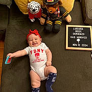 Luciana is registered to the contest to win money with this photo: announcement, baby, clothing, couch, dalmatian, firefighter_bear, floor, happy, headband, indoor, infant, letter_board, newborn, person, pillows, plush_toy, smile, socks, toy, wooden_floor