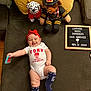 announcement, baby, clothing, couch, dalmatian, firefighter_bear, floor, happy, headband, indoor, infant, letter_board, newborn, person, pillows, plush_toy, smile, socks, toy, wooden_floor