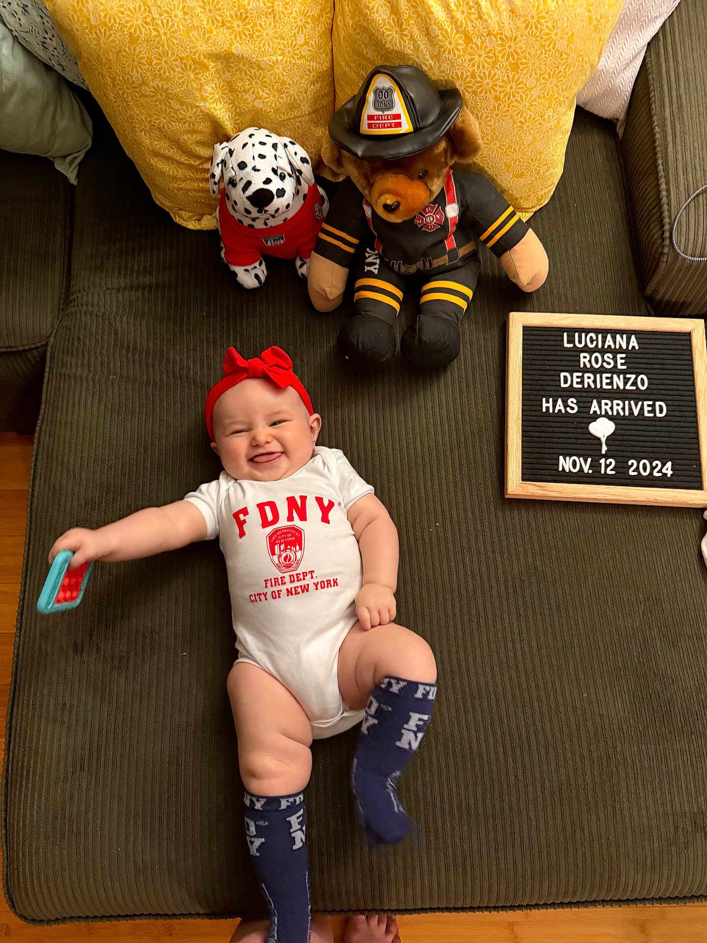 Luciana is registered to the contest to win money with this photo: announcement, baby, clothing, couch, dalmatian, firefighter_bear, floor, happy, headband, indoor, infant, letter_board, newborn, person, pillows, plush_toy, smile, socks, toy, wooden_floor