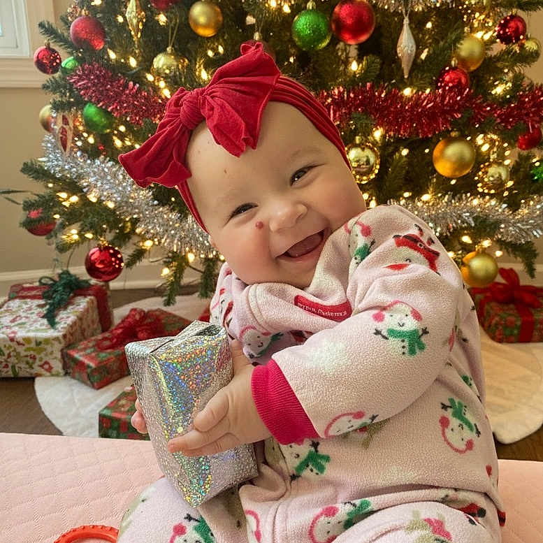 Norah is registered to the contest to win money with this photo: baby, child, christmas_tree, decorations, festive, floor, gift, happy, headband, holiday, holiday_season, indoors, lights, ornaments, pajamas, present, red_bow, smile, toy, wrapped_gift