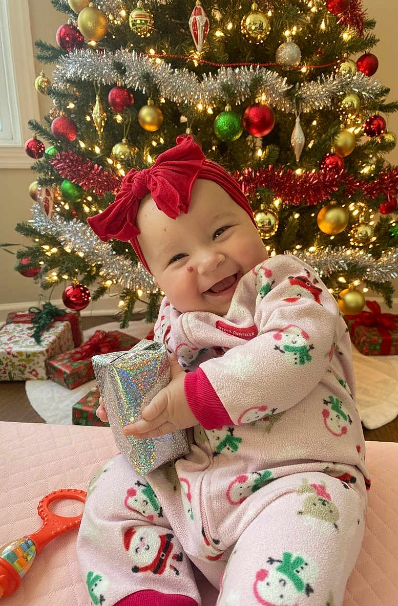 Norah is registered to the contest to win money with this photo: baby, child, christmas_tree, holiday, gift, pajamas, headband, red_bow, smile, happy, indoors, decorations, ornaments, lights, present, festive, floor, toy, wrapped_gift, holiday_season