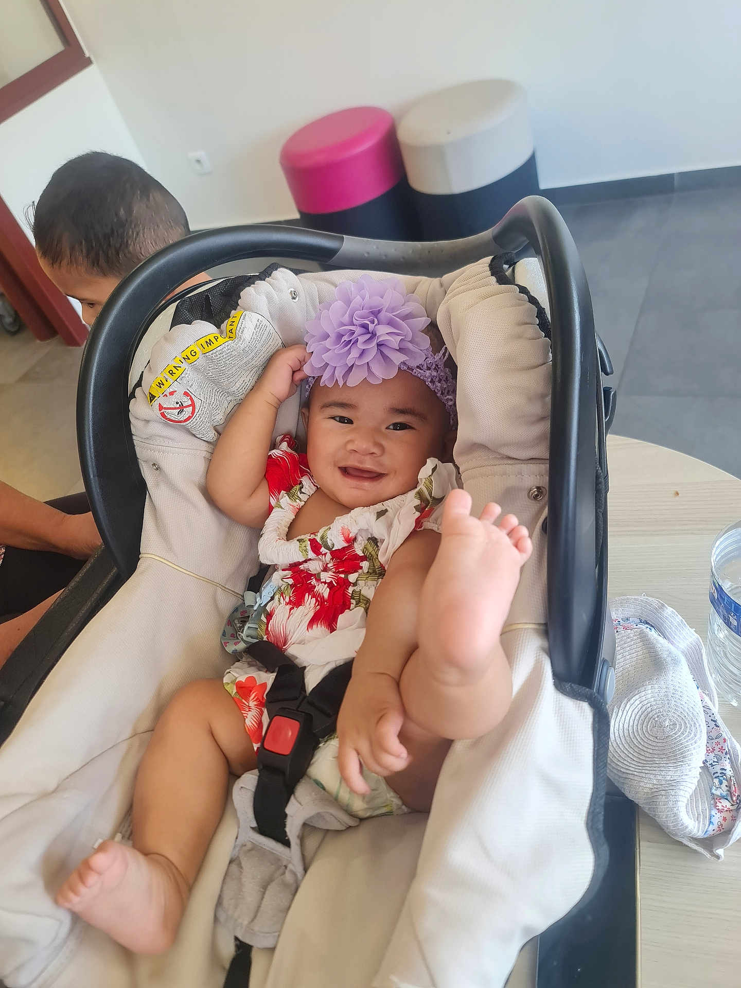 Tikena a rejoint le concours — aidez-le/la à gagner de superbes lots ! baby, baby_carrier, child, clothing, curious, floor, flower, foot, happy, headband, indoor, infant, person, playful, seat, smile, stool, table, toddler, water_bottle