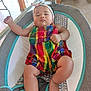arm, baby, baby_seat, child, chubby, colorful_clothing, cushion, cute, face, foot, headband, indoor, infant, infant_clothing, person, plaid, relaxed, resting, romper, skin