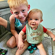 Caden is registered to the contest to win money with this photo: children, siblings, baby, boy, toddler, smiling, happy, portrait, indoor, home, carpet, couch, toy, playtime, blond_hair, blue_eyes, sitting, casual_clothing, bib, family