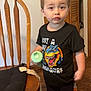 black_tshirt, casual_clothing, child, curious_expression, cushion, dinosaur_graphic, door, footwear, home_interior, indoor, orange_stripes, pants, person, short_hair, sippy_cup, standing, toddler, wooden_chair, wooden_floor, young_child