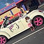 child, girl, car, convertible, hello_kitty, pink, rainbow, parking_lot, building, sign, smiling, outdoor, sunny, fashion, accessory, toy, vehicle, person, colorful, happy
