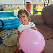Elena is registered to the contest to win money with this photo: toddler, balloon, smile, curly_hair, boots, white_shirt, inflatable_pool, hula_hoop, brown_couch, patio, outdoor, sunny, child, playful, happy, grass, backyard, fence, table, container