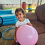 toddler, balloon, smile, curly_hair, boots, white_shirt, inflatable_pool, hula_hoop, brown_couch, patio, outdoor, sunny, child, playful, happy, grass, backyard, fence, table, container