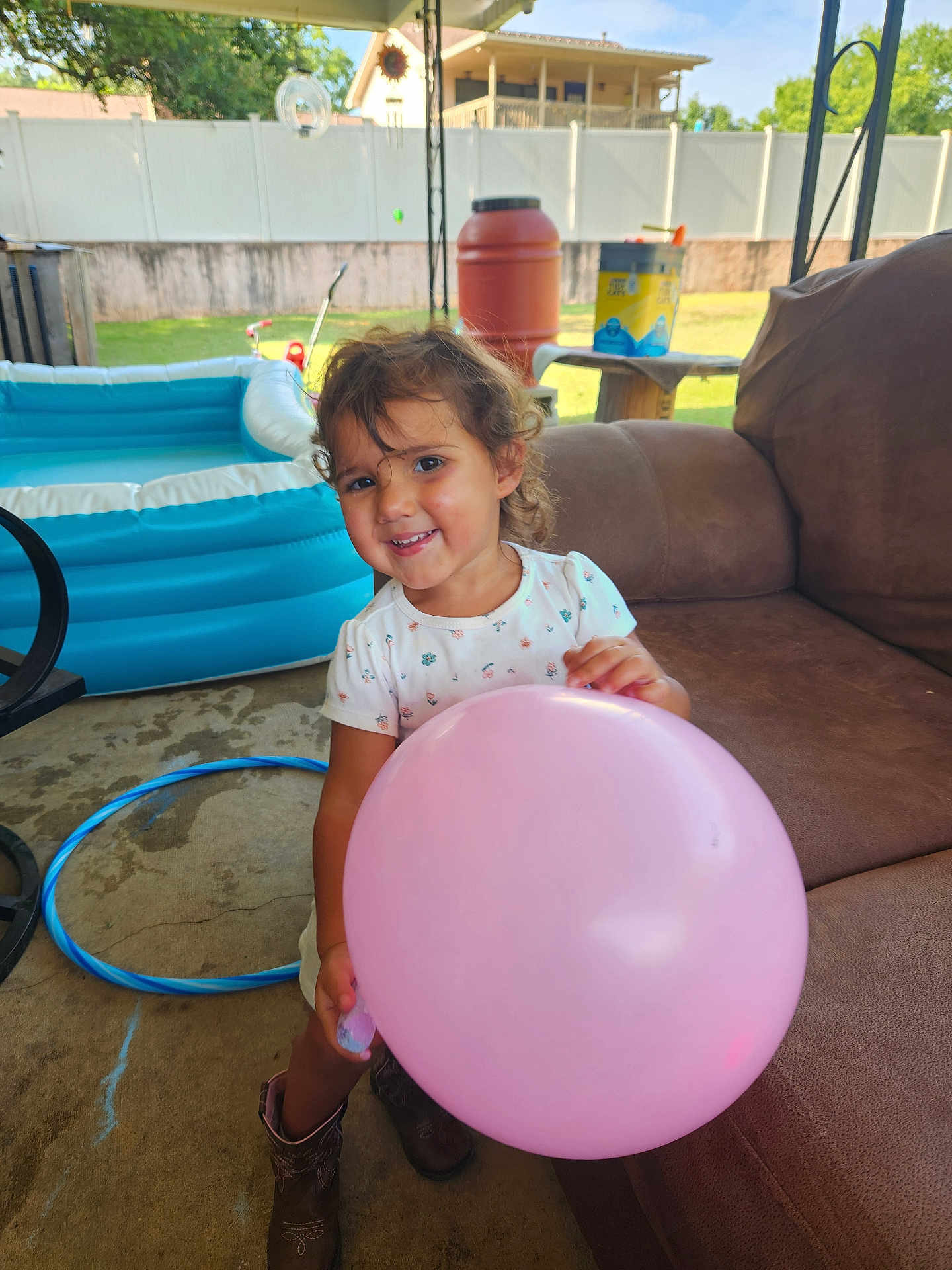 Elena is registered to the contest to win money with this photo: toddler, balloon, smile, curly_hair, boots, white_shirt, inflatable_pool, hula_hoop, brown_couch, patio, outdoor, sunny, child, playful, happy, grass, backyard, fence, table, container