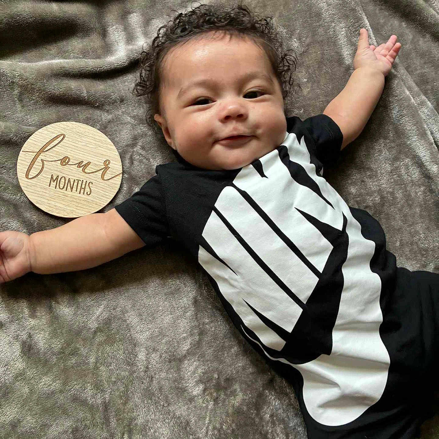 Zayden is registered to the contest to win money with this photo: arm, baby, black_clothing, blanket, cheeky, child, curly_hair, cute, face, four_months, happy, indoors, infant, lying_down, nike_logo, person, smile, soft_texture, wooden_disc, young