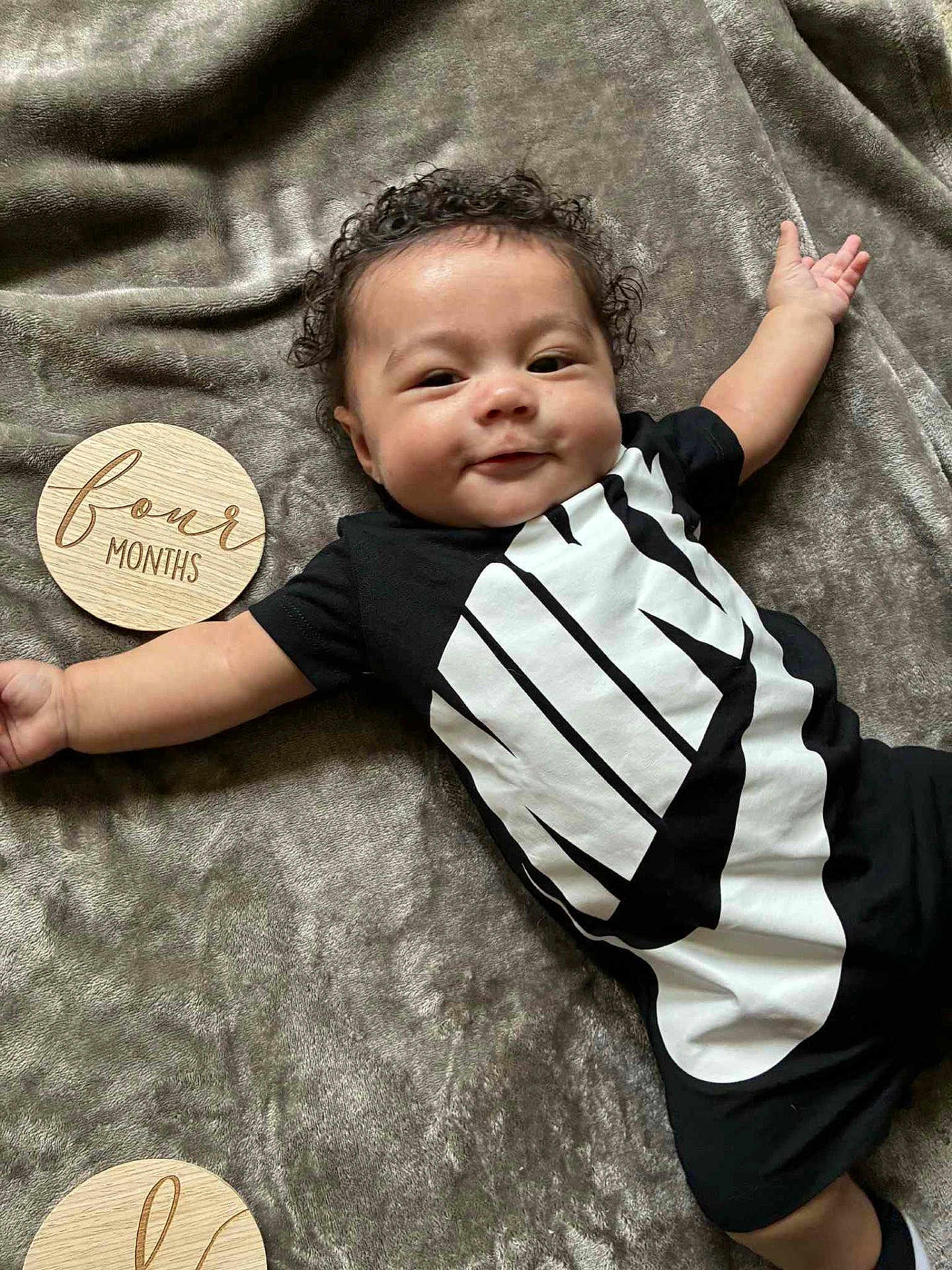 Zayden is registered to the contest to win money with this photo: baby, infant, smile, curly_hair, black_clothing, nike_logo, blanket, wooden_disc, four_months, lying_down, happy, face, arm, cheeky, indoors, soft_texture, child, cute, person, young