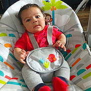Zayden is registered to the contest to win money with this photo: baby, infant, child, red_shirt, gray_pants, red_socks, baby_seat, colorful, animal_print, monkey, bird, straps, indoors, wooden_floor, curly_hair, cute, seated, looking_at_camera, playful, cozy