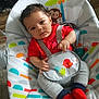 Zayden is registered to the contest to win money with this photo: baby, infant, child, red_shirt, gray_pants, red_socks, baby_bouncer, animal_print, monkey, bird, straps, indoor, carpet, wood_floor, hand, curly_hair, face, looking, seated, calm