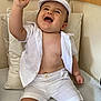 baby, child, laughing, white_clothing, hat, cushion, wooden_background, smiling, sitting, cute, happy, infant, shorts, shirt, footwear, indoors, portrait, raised_hand, playful, comfortable