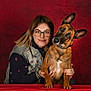 affection, animal, brown_dog, casual_clothing, companion, curious, denim_vest, dog, friendship, glasses, holding, indoor, looking, person, pet, portrait, red_background, smile, studio, woman