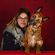 Olsa a rejoint le concours — aidez-le/la à gagner de superbes lots ! animal, brown_dog, casual_clothing, companionship, cute, denim_jacket, dog, friendly, glasses, hands, head_tilt, human, indoor, love, pet, portrait, red_background, smile, table, woman