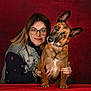 animal, brown_dog, casual_clothing, companionship, cute, denim_jacket, dog, friendly, glasses, hands, head_tilt, human, indoor, love, pet, portrait, red_background, smile, table, woman