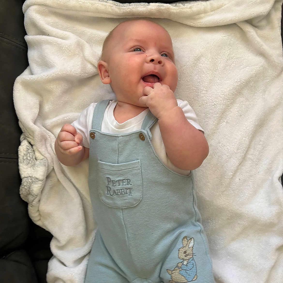 Gwylym is registered to the contest to win money with this photo: baby, blanket, child, clothing, comfort, couch, cute, face, fingers, hand, happy, indoor, infant, light_blue, lying_down, overalls, peter_rabbit, smiling, soft_texture, white