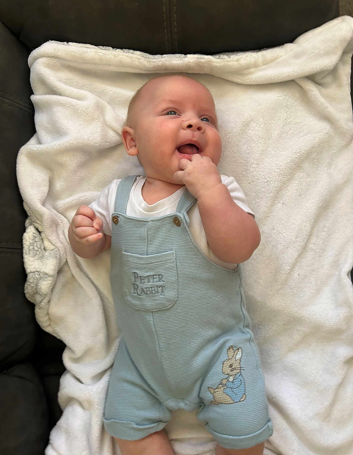 Gwylym is registered to the contest to win money with this photo: baby, infant, smiling, overalls, clothing, peter_rabbit, blanket, soft_texture, happy, cute, face, hand, fingers, lying_down, indoor, child, light_blue, white, comfort, couch