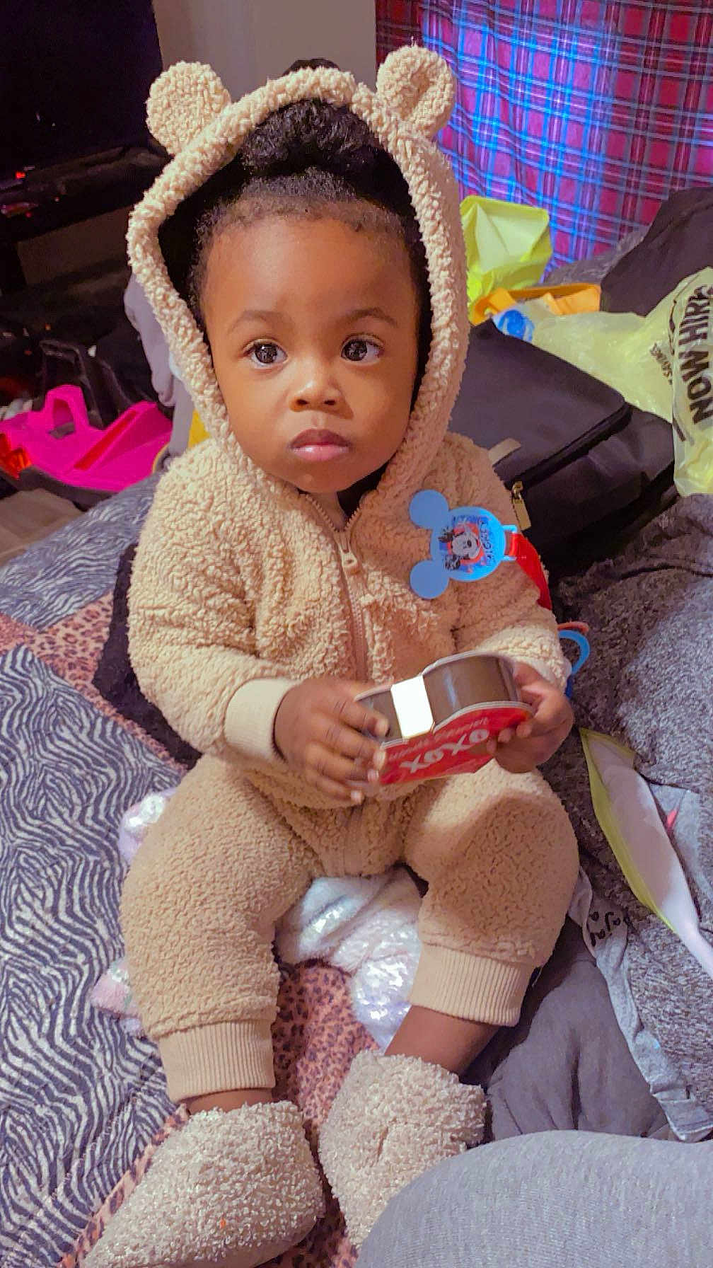 Jahmir is registered to the contest to win money with this photo: toddler, child, onesie, bear_ears, plush_clothing, indoor, bed, blanket, toy, heart_shape, xoxo, patterned_bedspread, cozy, sitting, person, cute, warm_clothing, expression, holding, footwear