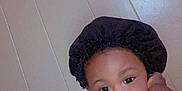 Jahmir is registered to the contest to win money with this photo: baby, bonnet, brown_skin, casual, child, curious, cute, expression, face, hand, headwear, indoors, person, portrait, room, sitting, toddler, wall, white_clothing, young_child