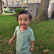 Lucas is registered to the contest to win money with this photo: child, toddler, smiling, grass, tree, urban, park, shorts, tshirt, sandals, green_clothing, happy, outdoor, daylight, building, sidewalk, nature, person, cute, young