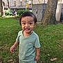 child, toddler, smiling, grass, tree, urban, park, shorts, tshirt, sandals, green_clothing, happy, outdoor, daylight, building, sidewalk, nature, person, cute, young