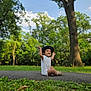Lucas joined the competition — help win amazing prizes! toddler, child, hat, white_onesie, smiling, sitting, arms_up, outdoor, park, grass, trees, path, nature, daylight, happy, footwear, young_child, greenery, sky, casual