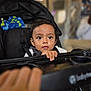 toddler, child, stroller, baby, curious, indoor, hand, face, black_hair, clothing, seat, grip, tray, person, infant, portrait, expression, blurred_background, cute, young