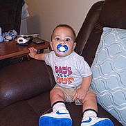 Malachi is registered to the contest to win money with this photo: baby, pacifier, sneakers, socks, shirt, shorts, couch, pillow, table, remote_control, phone, armrest, living_room, child, person, indoor, furniture, curious, seated, wide_eyes