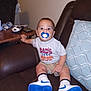 baby, pacifier, sneakers, socks, shirt, shorts, couch, pillow, table, remote_control, phone, armrest, living_room, child, person, indoor, furniture, curious, seated, wide_eyes