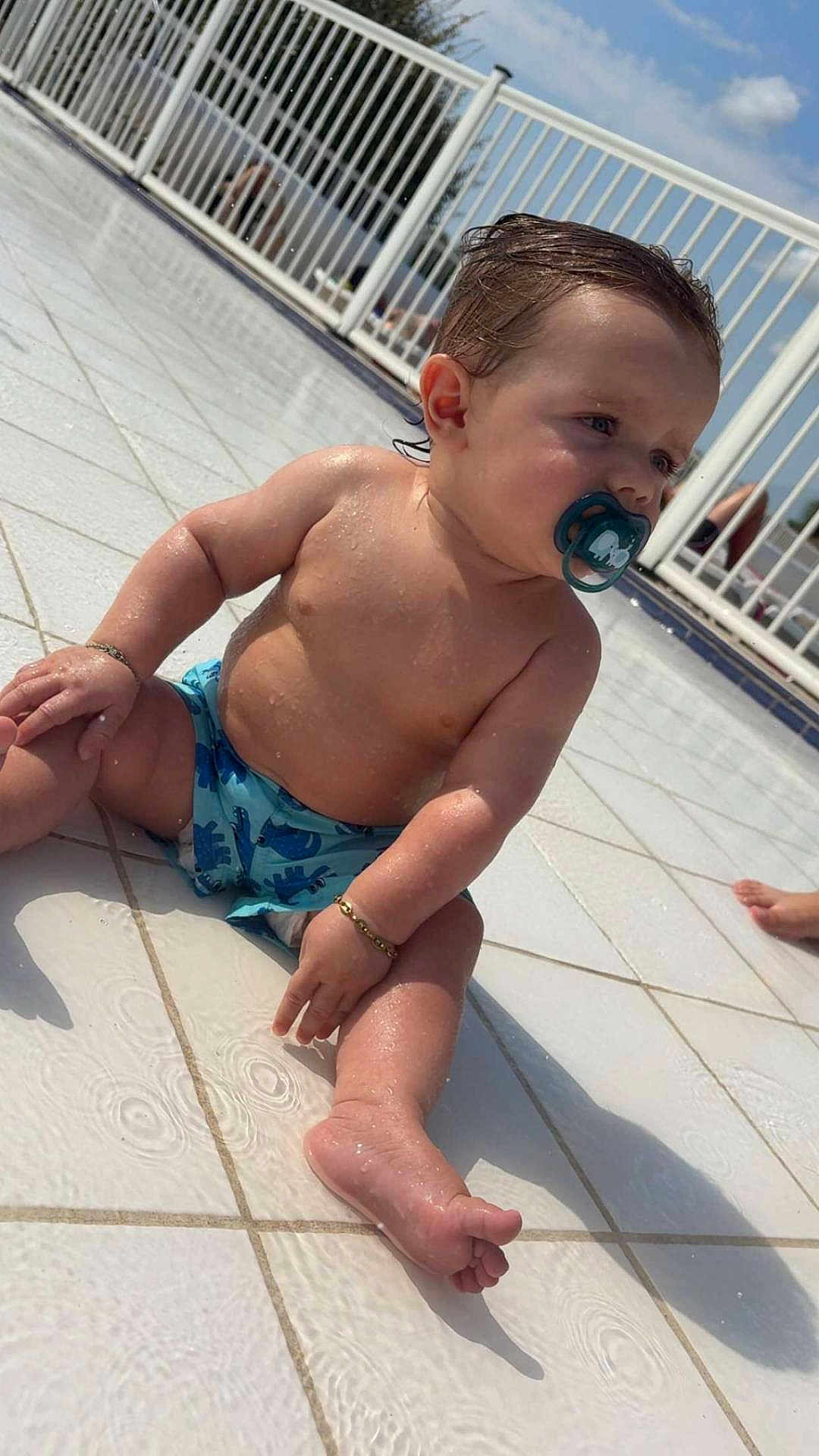 Lenzo participe au concours pour gagner de l'argent avec cette photo : baby, child, pacifier, water, wet_skin, poolside, tiled_floor, sunlight, shadow, bracelet, blue_swim_diaper, outdoor, summer, sitting, cute, infant, skin, foot, hand, face