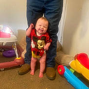 Mia joined the competition — help win amazing prizes! baby, smiling_child, infant, mickey_mouse_shirt, standing_with_support, adult_legs, jeans, boots, toys, plastic_toy, carpet, indoor, high_chair, walking_assist, happy, playroom, cute, red_clothing, bare_feet, laughing