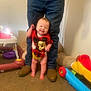 baby, smiling_child, infant, mickey_mouse_shirt, standing_with_support, adult_legs, jeans, boots, toys, plastic_toy, carpet, indoor, high_chair, walking_assist, happy, playroom, cute, red_clothing, bare_feet, laughing