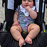 baby, infant, child, stroller, sitting, smiling, portrait, outdoor, pavement, canopy, harness, floral_clothing, bib, bare_feet, toes, black_hair, face, safety_labels, happy, photography