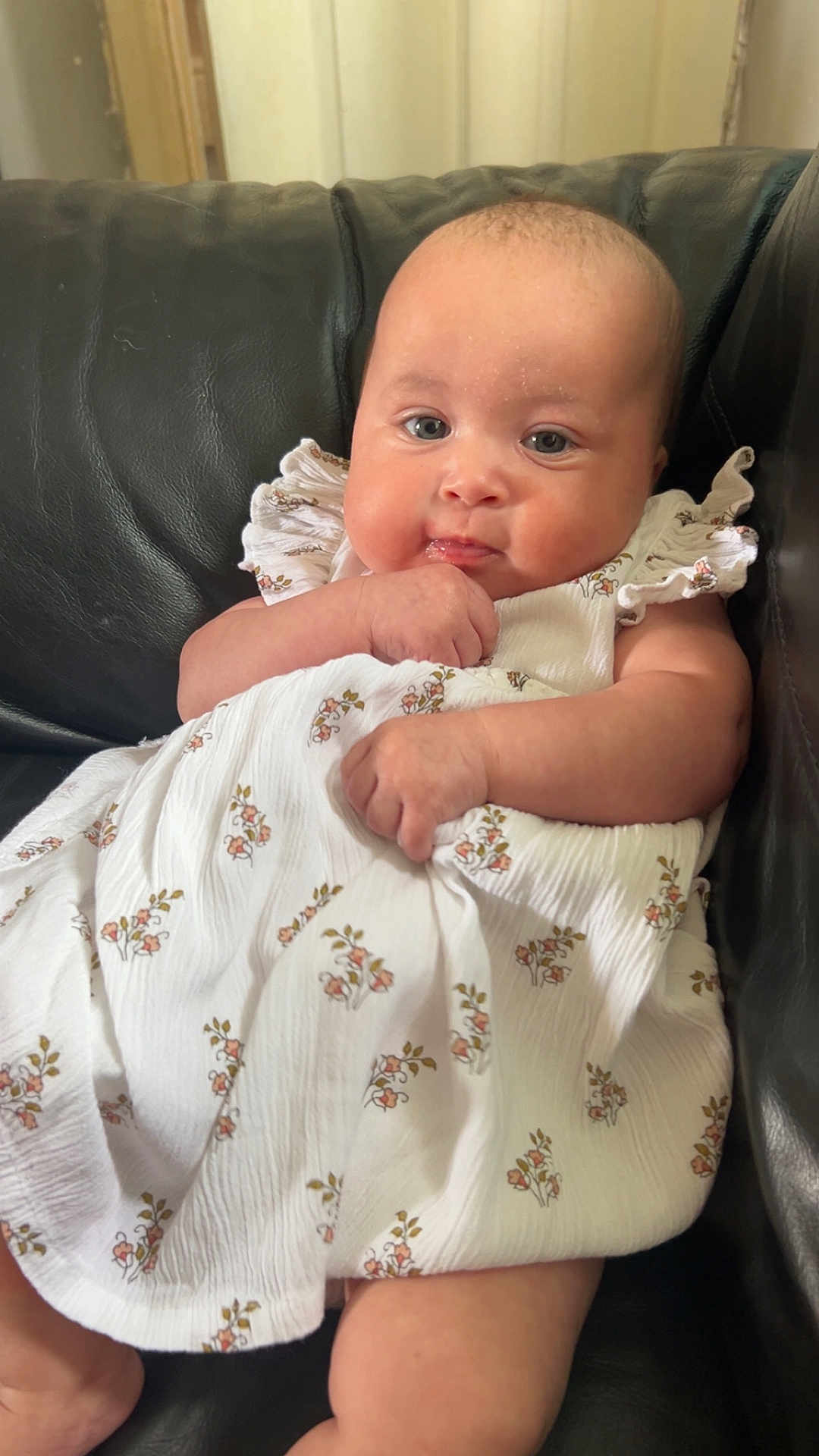 Gracie-May is registered to the contest to win money with this photo: baby, child, infant, dress, white_dress, floral_pattern, couch, leather, black_couch, sitting, hand, face, cheeks, expression, indoor, portrait, young_child, cute, skin, person