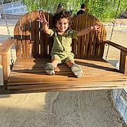 Ethan Perez is registered to the contest to win money with this photo: child, toddler, swing, wooden_swing, outdoor, sunlight, greenery, tree, happy, curly_hair, casual_clothing, sand, playground, daylight, smiling, person, nature, bench, arm_stretch, fun