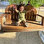child, toddler, swing, wooden_swing, outdoor, sunlight, greenery, tree, happy, curly_hair, casual_clothing, sand, playground, daylight, smiling, person, nature, bench, arm_stretch, fun