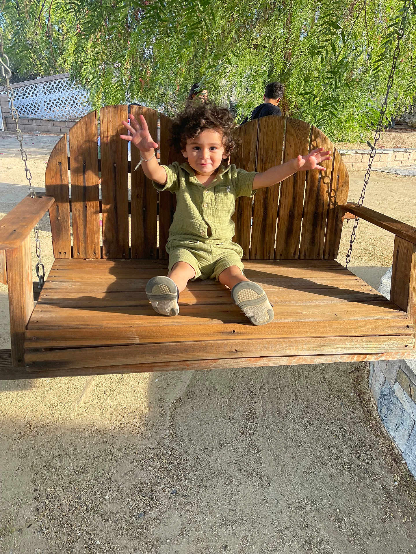 Ethan Perez is registered to the contest to win money with this photo: child, toddler, swing, wooden_swing, outdoor, sunlight, greenery, tree, happy, curly_hair, casual_clothing, sand, playground, daylight, smiling, person, nature, bench, arm_stretch, fun