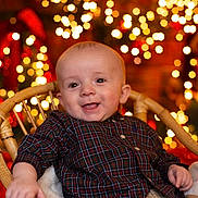 Lennie joined the competition — help win amazing prizes! baby, child, plaid_shirt, wicker_chair, holiday_lights, bokeh, smiling, portrait, indoor, festive, warm_lighting, cute, happy, infant, cozy, red, green, beige, background, celebration