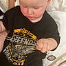 baby, child, toddler, sleepy, sitting, bed, bedding, black_shirt, graphic_shirt, chain, toy, hands, face, portrait, indoor, bedroom, adult_arm, watch, blanket, cozy