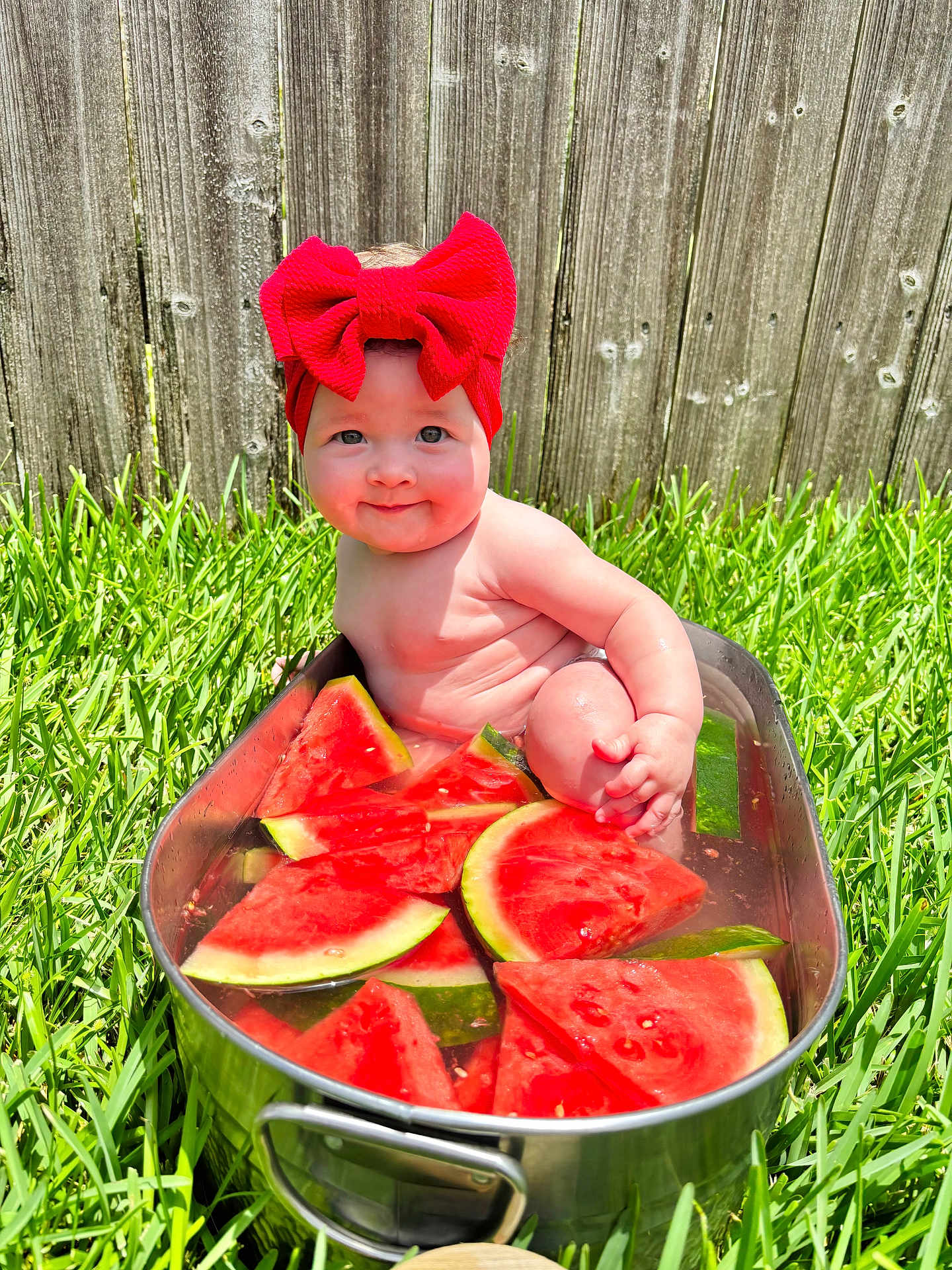 Avalyn is registered to the contest to win money with this photo: baby, child, red_bow, watermelon, water, metal_tub, grass, outdoor, summer, cute, headband, smiling, fence, greenery, sitting, chubby_cheeks, sunlight, playful, fruit, skin