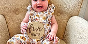 Avalyn is registered to the contest to win money with this photo: baby, child, smiling, headband, yellow_bow, floral_outfit, armchair, beige, wooden_sign, five_months, happy, sitting, cute, infant, portrait, indoors, texture, person, feet, hand