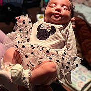 Kailynn is registered to the contest to win money with this photo: newborn, baby, infant, child, portrait, smiling, face, eyes_closed, outfit, dress, polka_dots, bow, mouse_design, held, adult_hands, indoor, bedroom, blanket, soft_lighting, family